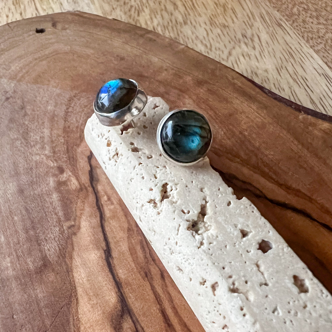 A pair of sterling silver earrings with labradorite studs placed on a beige textured surface.