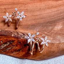 Load image into Gallery viewer, Rose gold earrings with three flower motifs and a central star, set with clear marquise-cut stones, displayed on a textured brown wood background.
