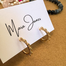 Load image into Gallery viewer, A pair of gold-plated detailed earring hoops displayed in front of a business card with the name &#39;Mara James&#39;.
