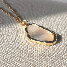 Load image into Gallery viewer, A gold-plated necklace with an oval-shaped pendant containing a white organic shell, displayed on a beige textured surface.
