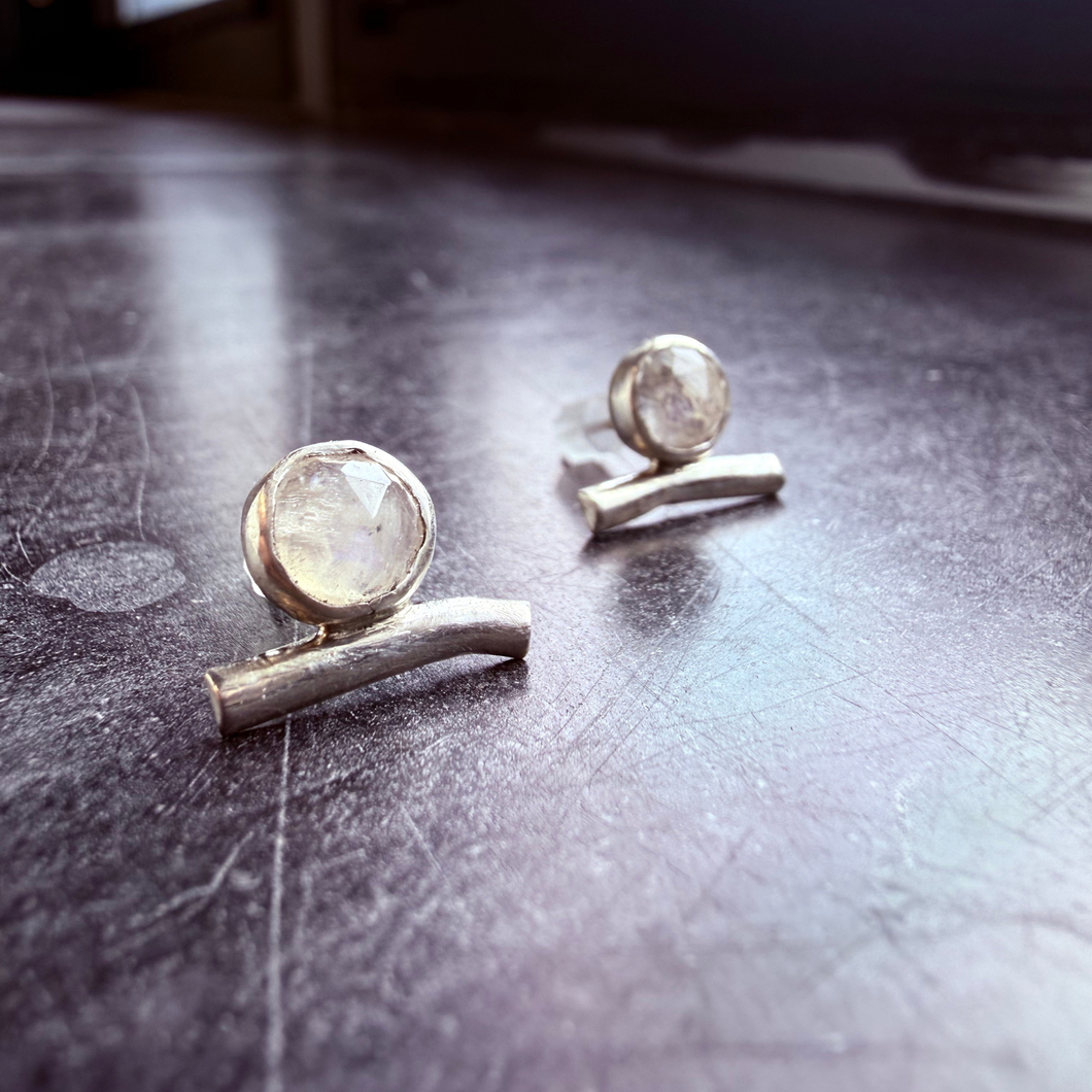 Handcrafted sterling silver earrings featuring faceted rainbow moonstones set above curved silver bars, photographed on a dark textured background.
