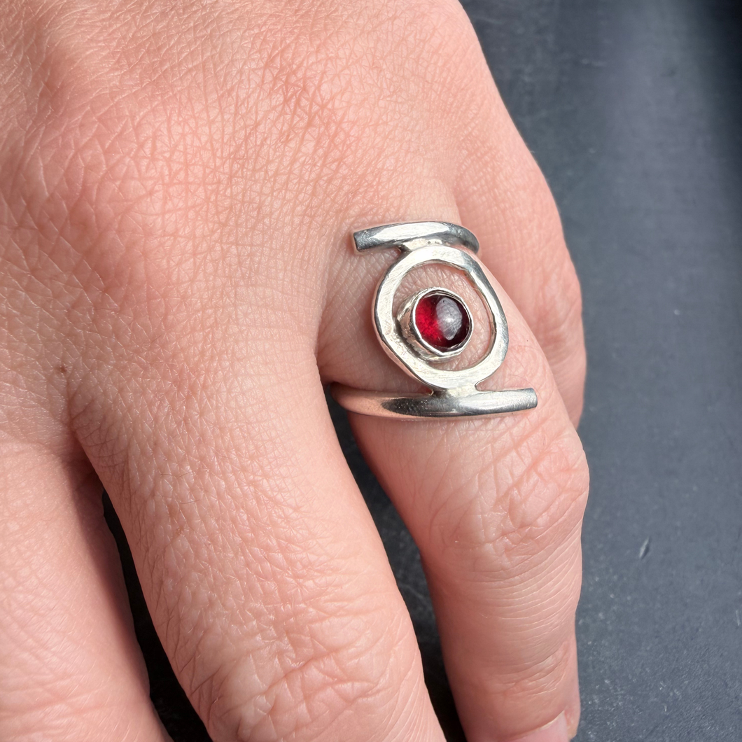 Handcrafted sterling silver ring featuring a round red garnet cabochon set in an open circular design, worn on hand against a dark background.