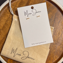 Load image into Gallery viewer, A pair of gold-plated pave bar earrings on a white card with the brand name &#39;Mara James&#39; written below, presented on a wooden surface.
