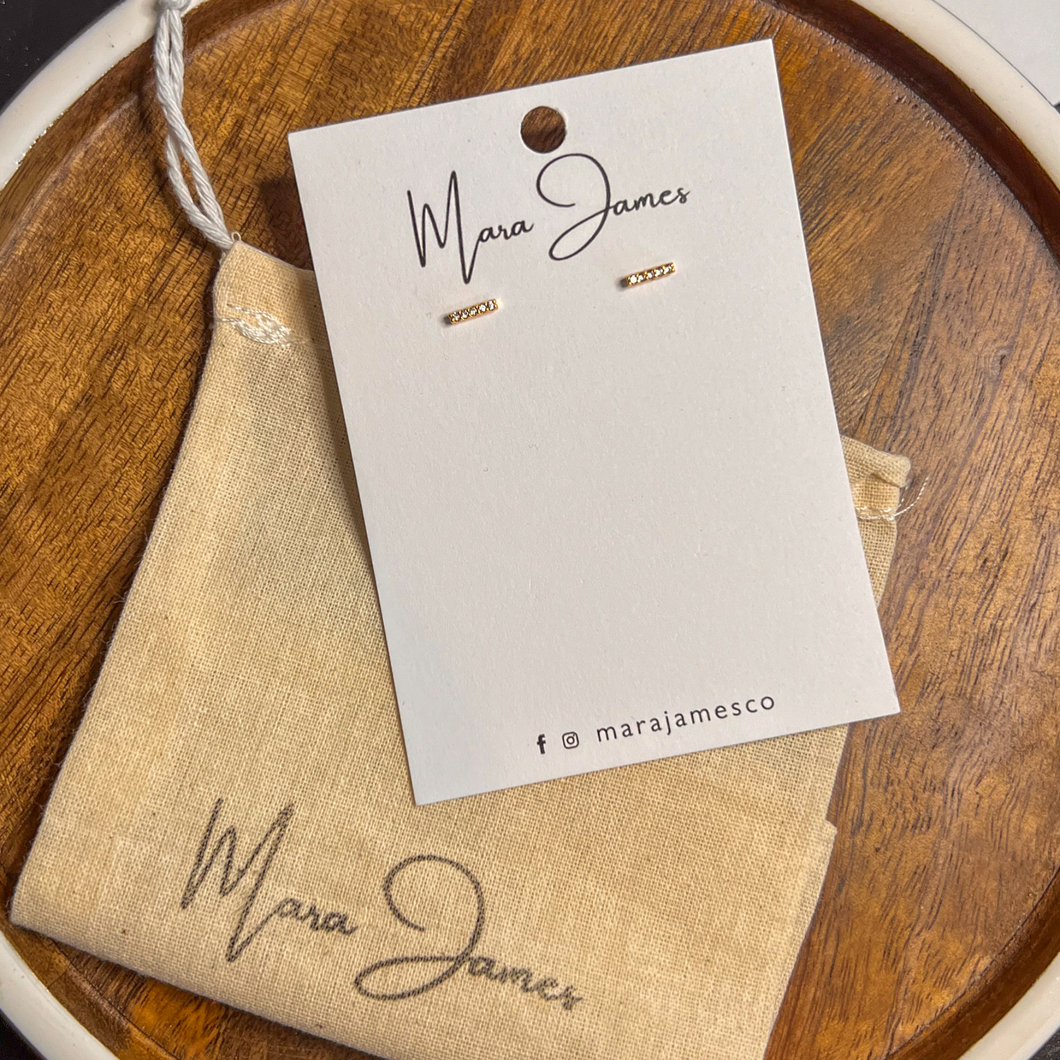 A pair of gold-plated pave bar earrings on a white card with the brand name 'Mara James' written below, presented on a wooden surface.