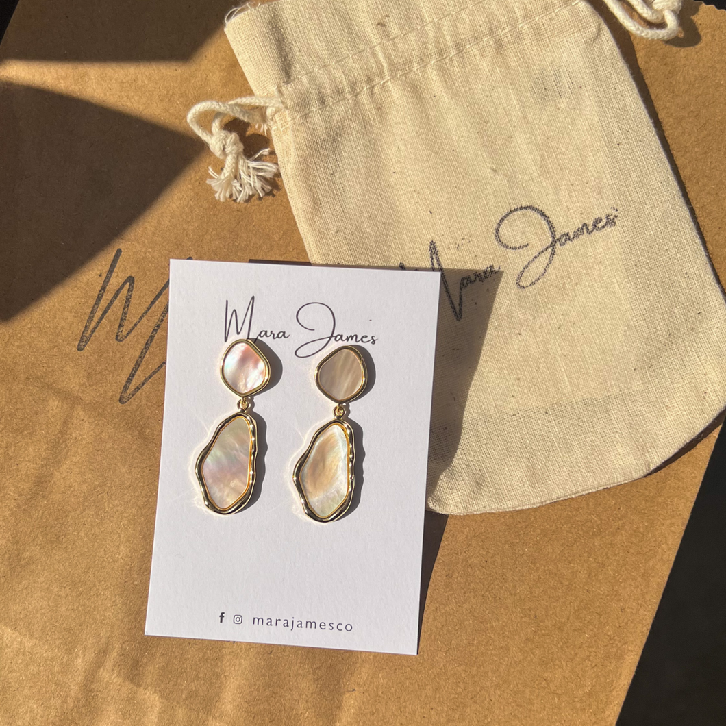A pair of gold-plated shell earrings displayed on a branded card with the name 'Mara James'. The earrings are crafted with a unique organic shape and are dangling.