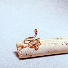 Load image into Gallery viewer, A gold snake ring with a small clear gemstone on its head, displayed on a textured beige stone block against a light gray background.
