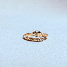 Load image into Gallery viewer, A gold snake ring with a textured scale pattern and a small clear gemstone on its head, resting on a light gray textured surface.
