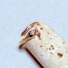 Load image into Gallery viewer, A gold snake ring with a small clear gemstone on its head, displayed on a textured beige stone cylinder against a light gray background.
